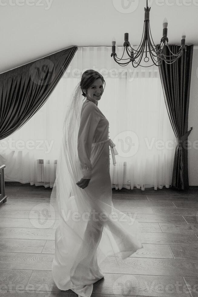 A woman in a white dress is standing in front of a window. She is wearing a veil and she is happy photo