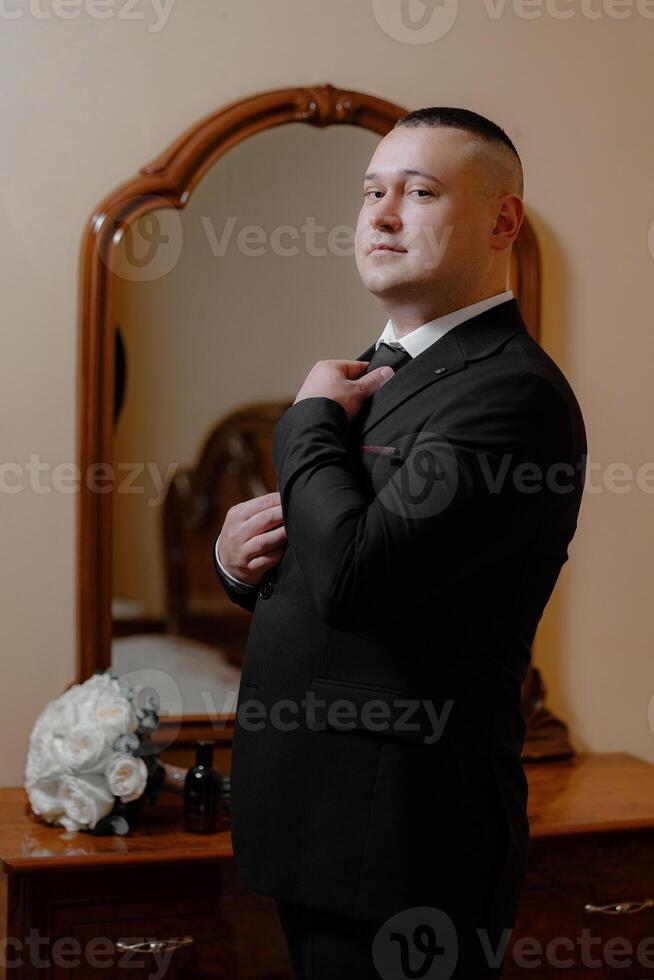 A man in a suit is standing in front of a mirror, adjusting his tie. Concept of formality and attention to detail, as the man takes the time to ensure his appearance is polished and professional photo