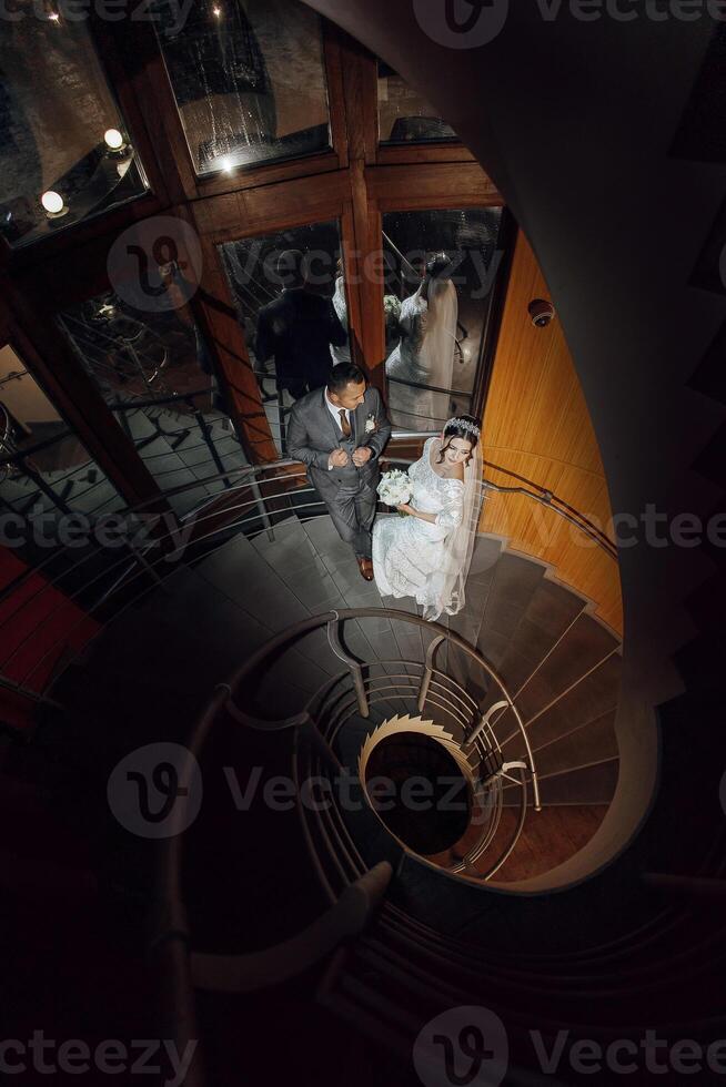 A bride and groom are standing on a spiral staircase. The bride is wearing a white dress and the groom is wearing a suit. They are both smiling and looking at the camera photo
