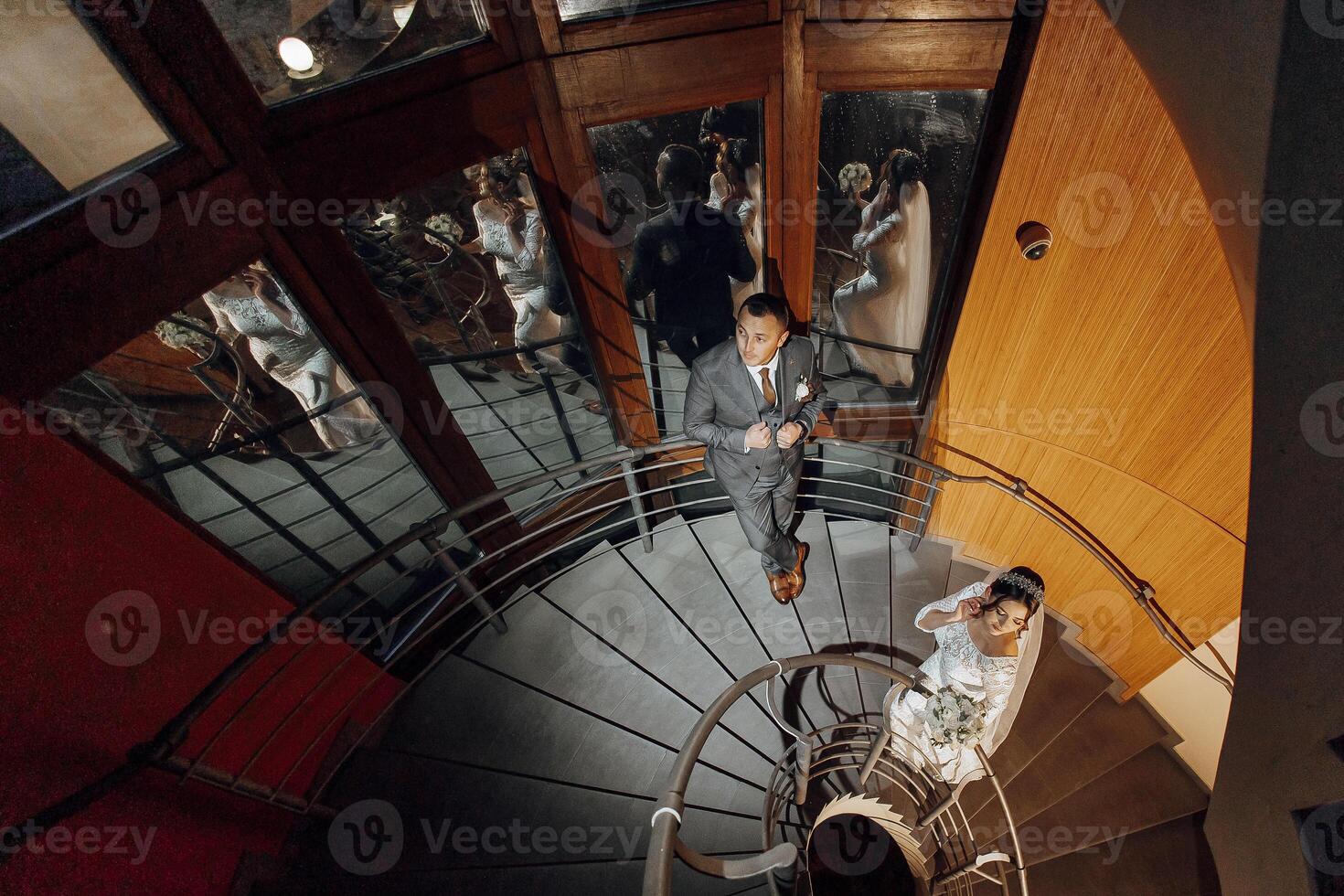 A bride and groom are walking down a spiral staircase. The bride is wearing a white dress and the groom is wearing a suit. The scene is set in a large room with many people in the background photo