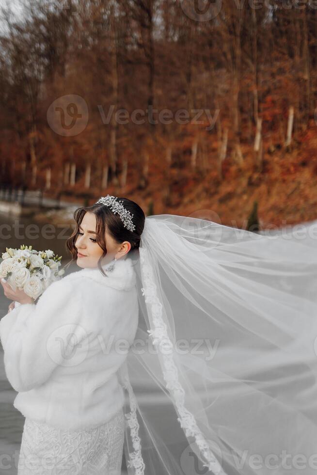 A woman in a white jacket is holding a bouquet of flowers. She is wearing a veil and a headband. The scene is set in a forest with trees in the background photo