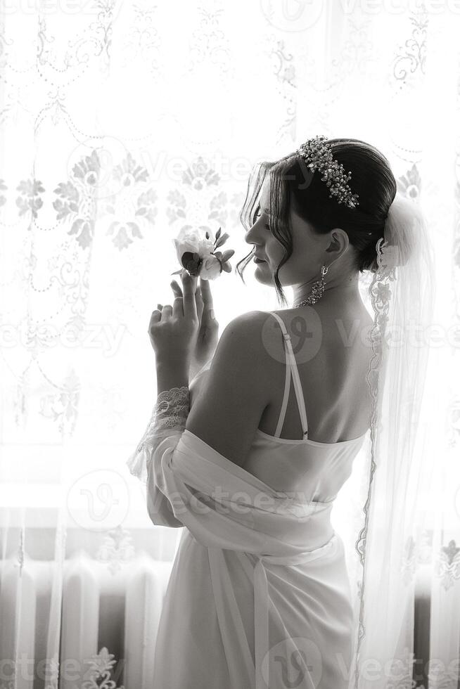 A woman in a wedding dress holding a flower. The image has a romantic and elegant mood. The woman is wearing a veil and a necklace, and she is standing in front of a window photo