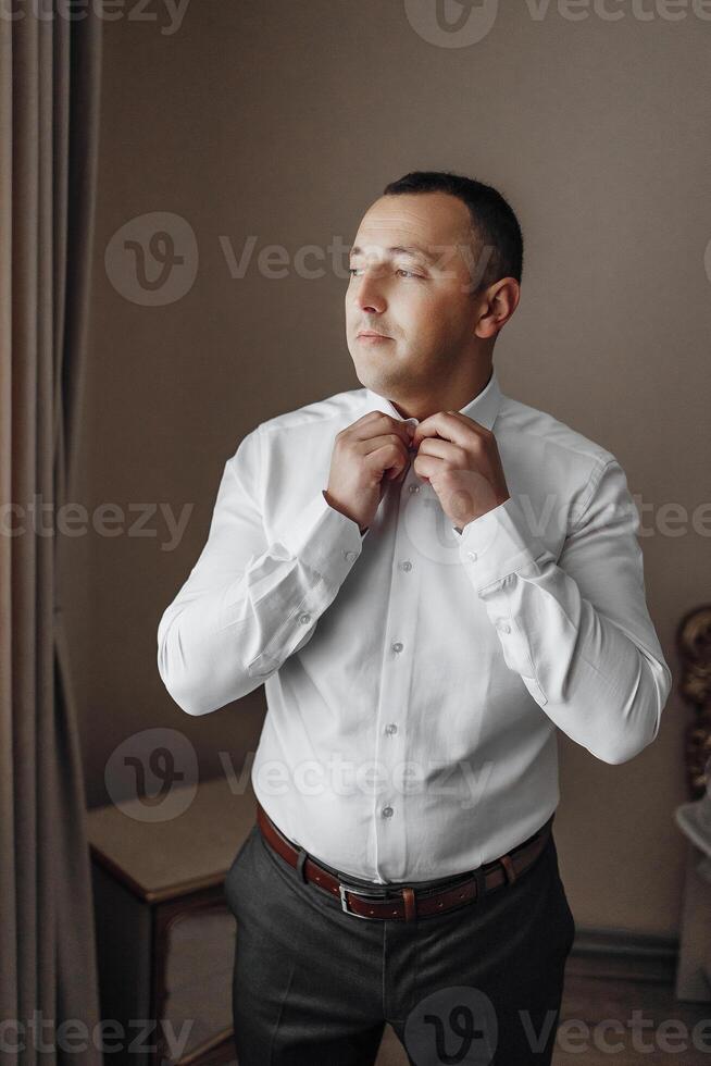 A man is getting ready to dress up in a white shirt and gray pants. He is adjusting his tie and looking out the window photo