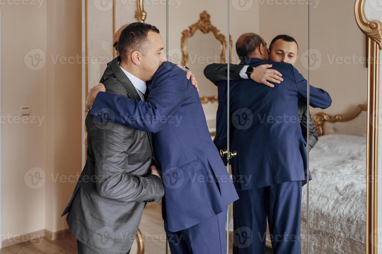Two men hug each other in a room with a mirror. Scene is warm and friendly photo
