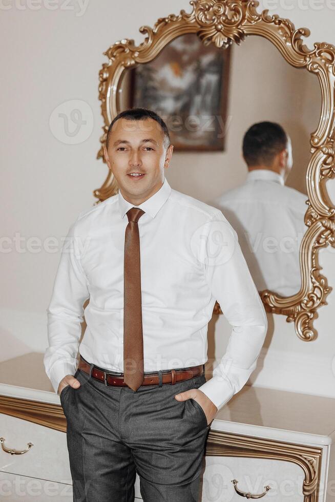 A man in a suit and tie stands in front of a mirror. He is wearing a brown tie and a brown belt photo