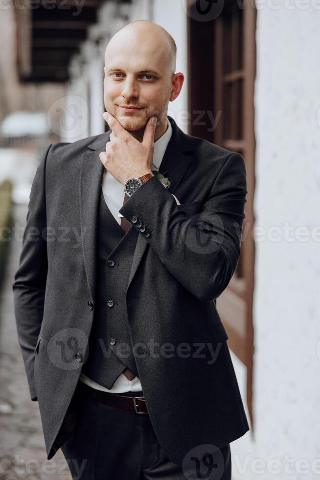 A man in a suit and vest is standing in front of a building. He is wearing a watch and a green flower in his hand. Concept of formality and elegance, as the man is dressed in a suit photo