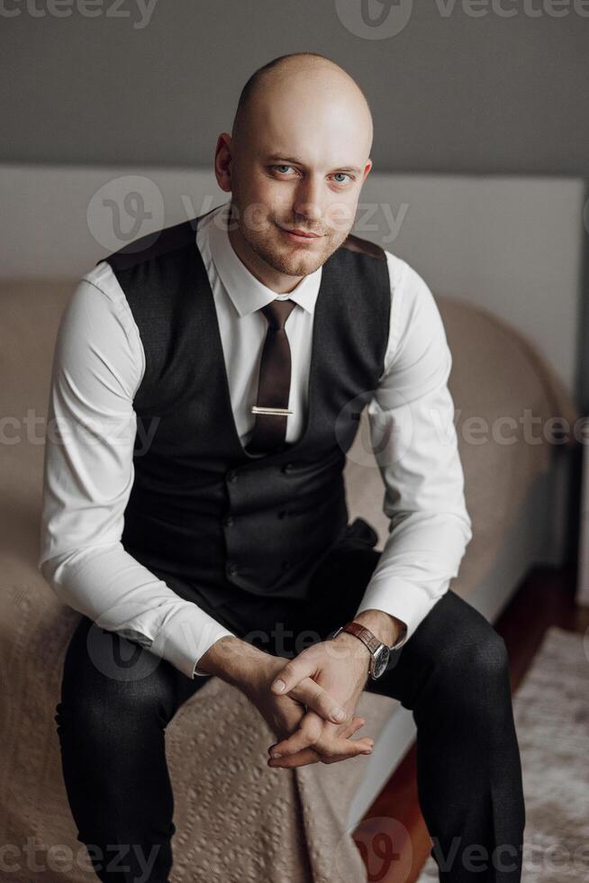 A man in a suit and vest sits on a bed. He is smiling and wearing a tie photo