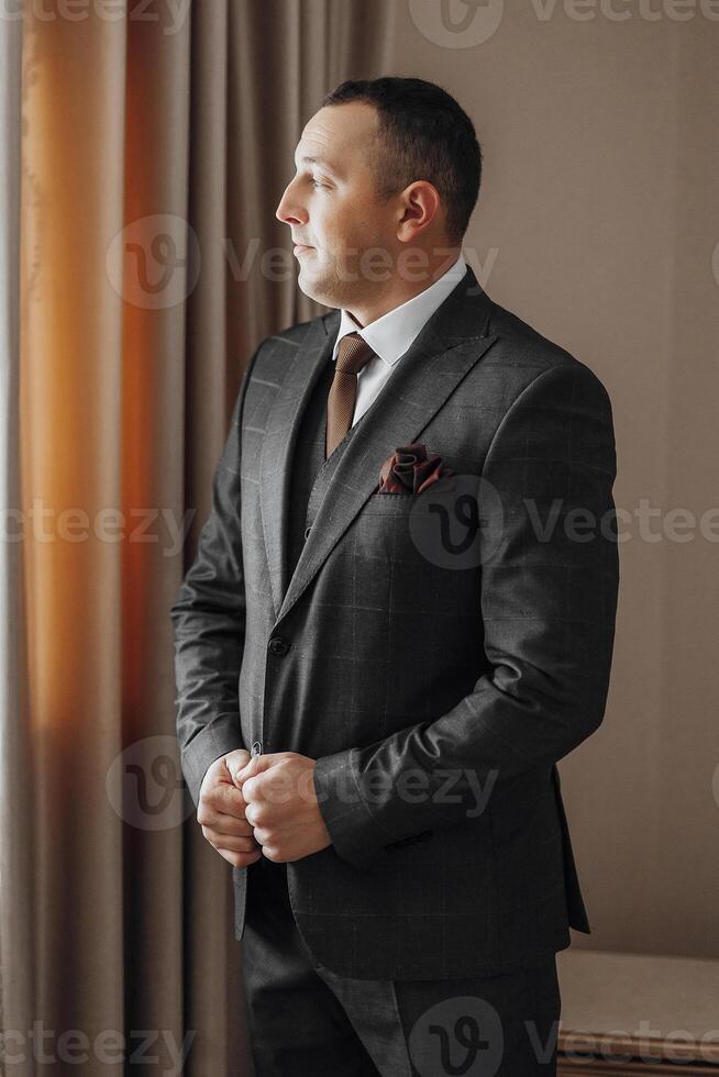 A man in a suit and tie stands in front of a window. He is wearing a red rose tie and a black suit photo