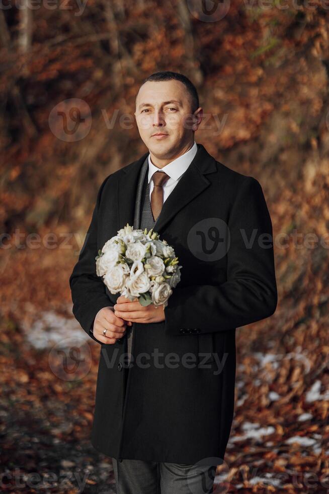A man wearing a black coat and tie holding a bouquet of flowers. The man is standing in a forest with a backdrop of trees photo