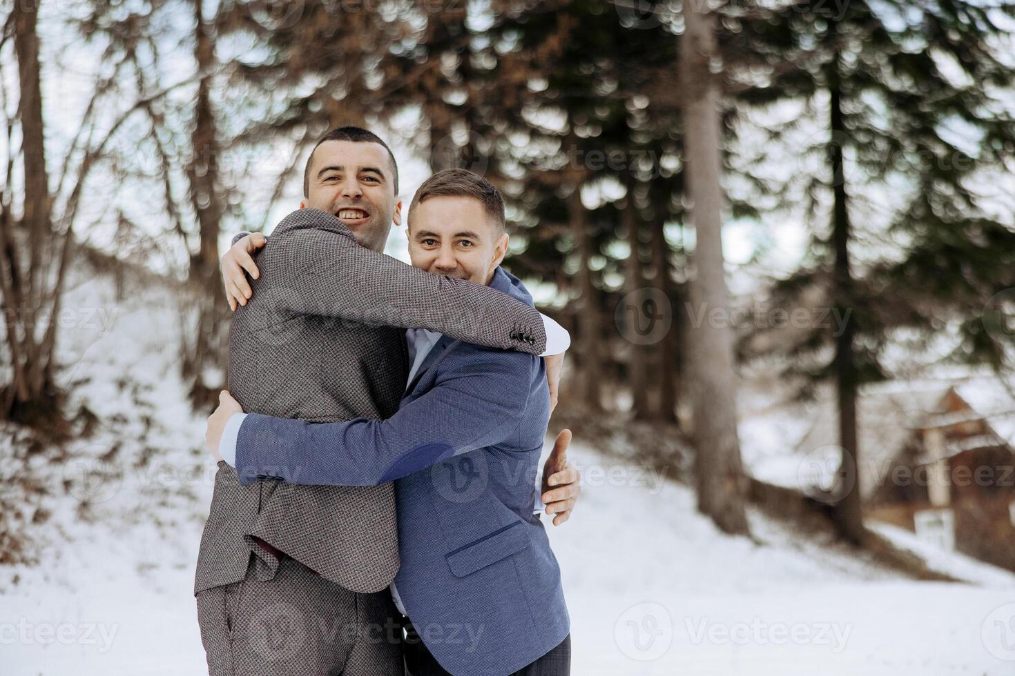 Two men hug each other in the snow. One of them is wearing a blue jacket. Scene is warm and friendly photo