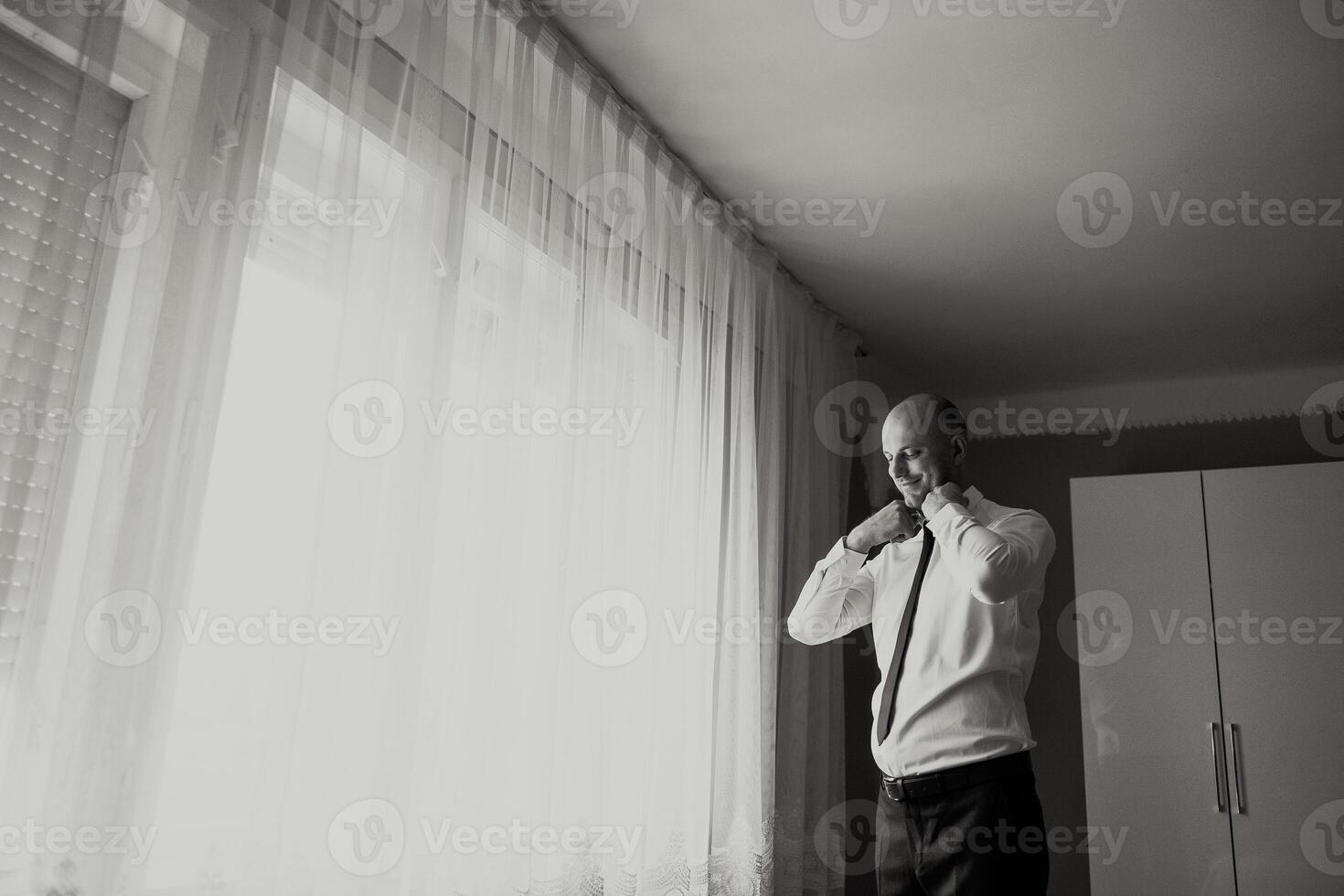 A man is getting ready for work, putting on his tie and adjusting his collar. The room is dimly lit, with a window letting in a small amount of light photo