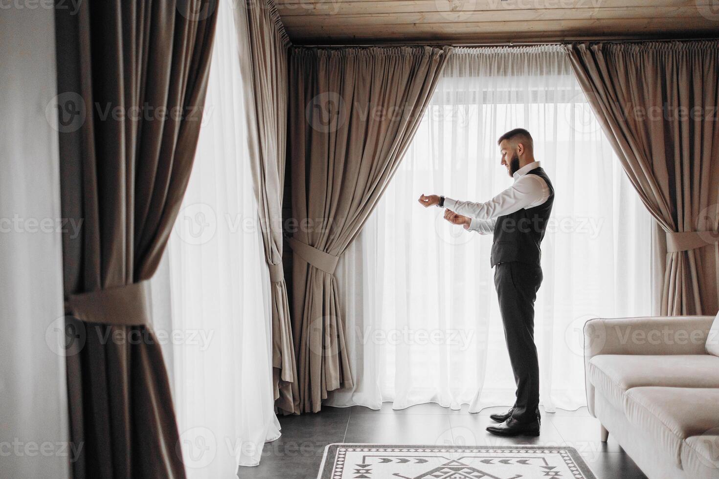 A man in a suit is standing in front of a window, adjusting his tie. The room is decorated with curtains and a rug, giving it a cozy and comfortable atmosphere photo
