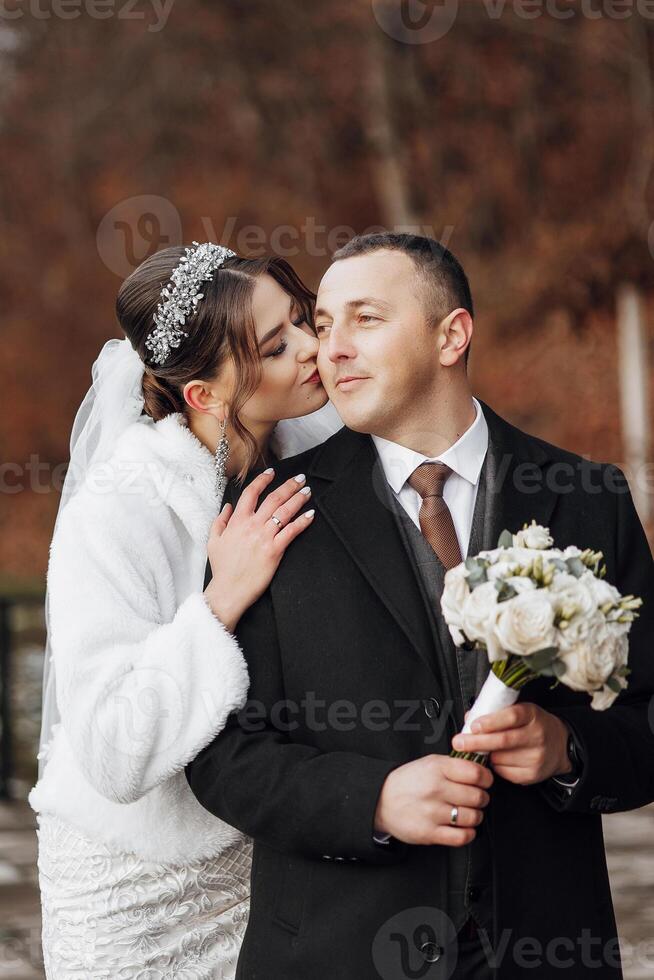 A bride and groom are kissing each other's cheeks. The bride is wearing a white coat and the groom is wearing a black suit. The groom is holding a bouquet of flowers photo