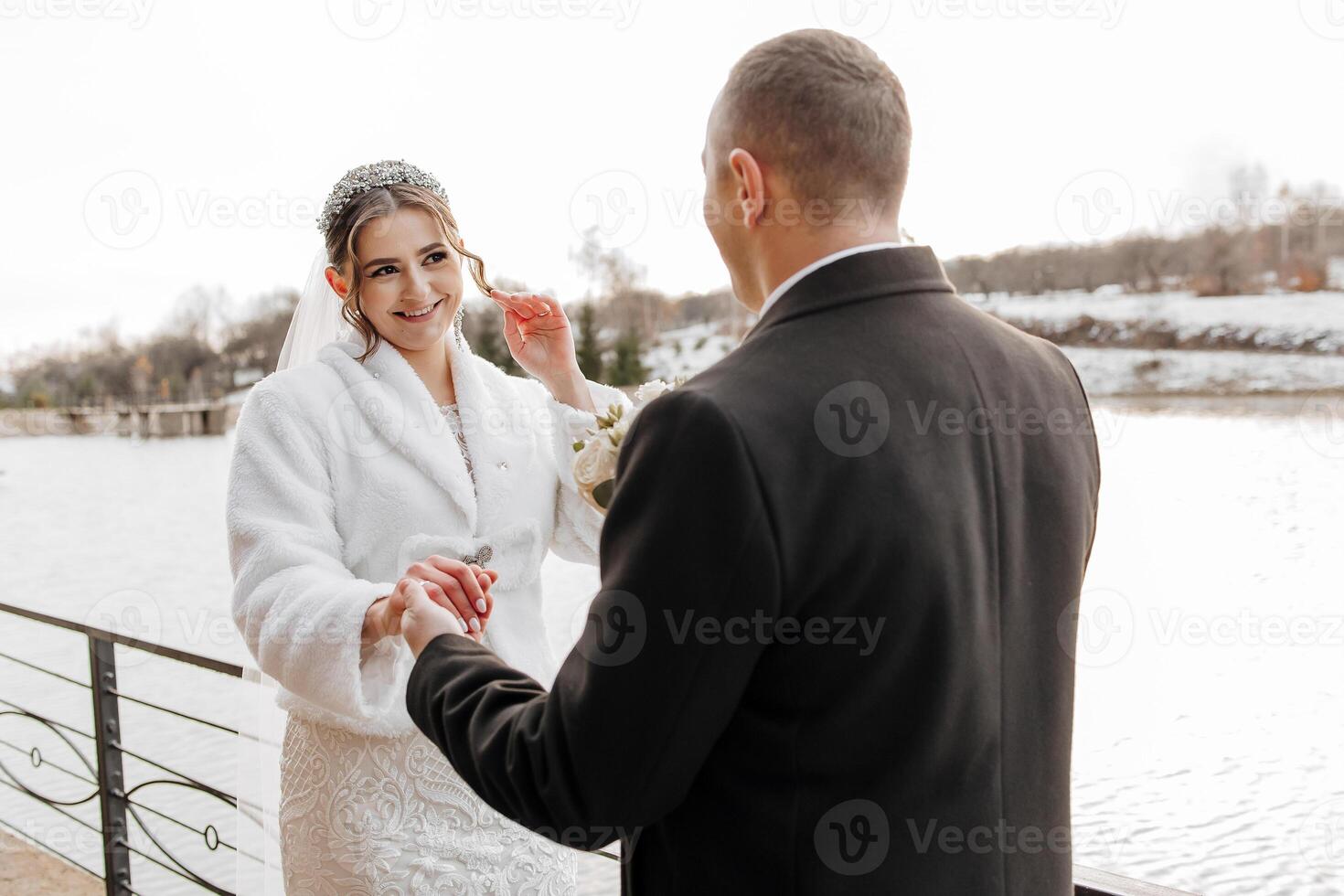 A bride and groom are standing on a bridge near a body of water. The bride is wearing a white coat and the groom is wearing a black suit. They are holding hands and smiling at the camera photo