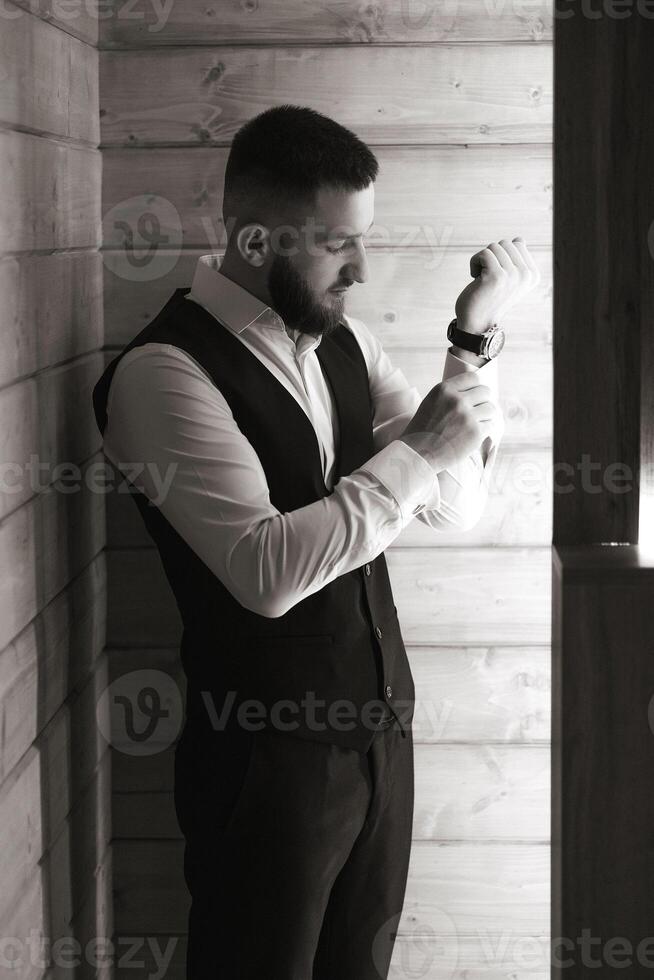 A man in a black vest is getting dressed. He is wearing a watch and has a beard photo