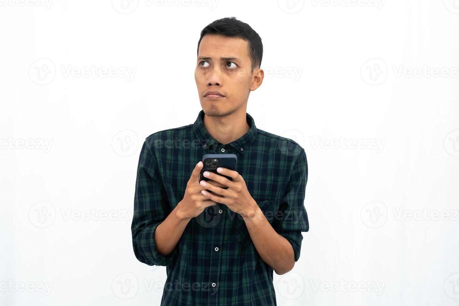 Asian man wearing a green checkered casual shirt is thinking while holding a smartphone. Isolated white background. photo