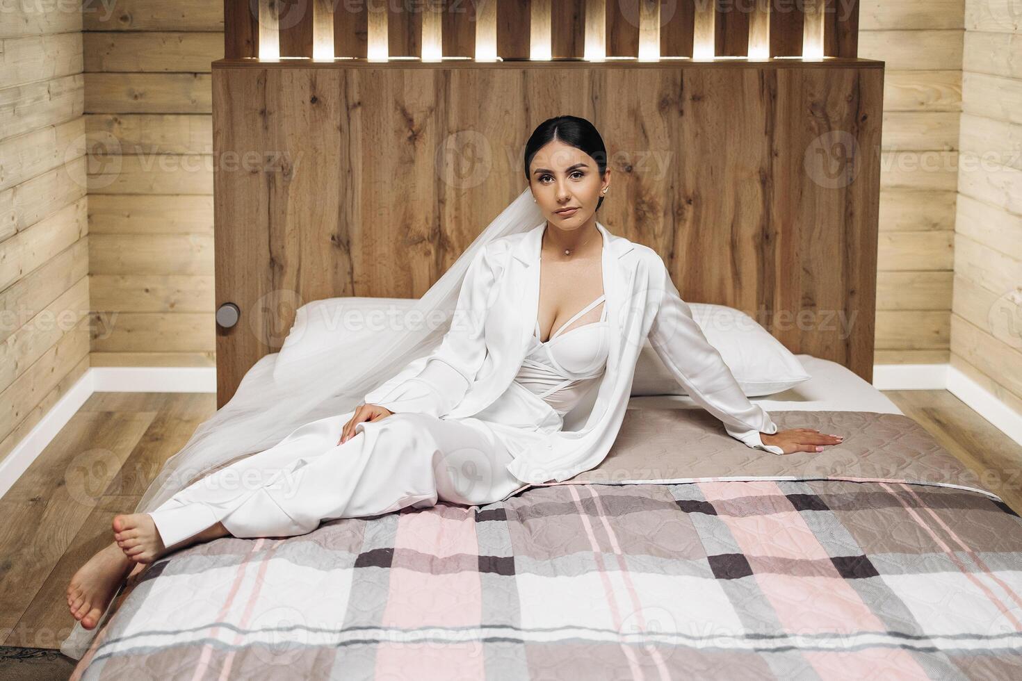 A woman is sitting on a bed in a room with a wooden headboard. She is wearing a white dress and a white jacket photo