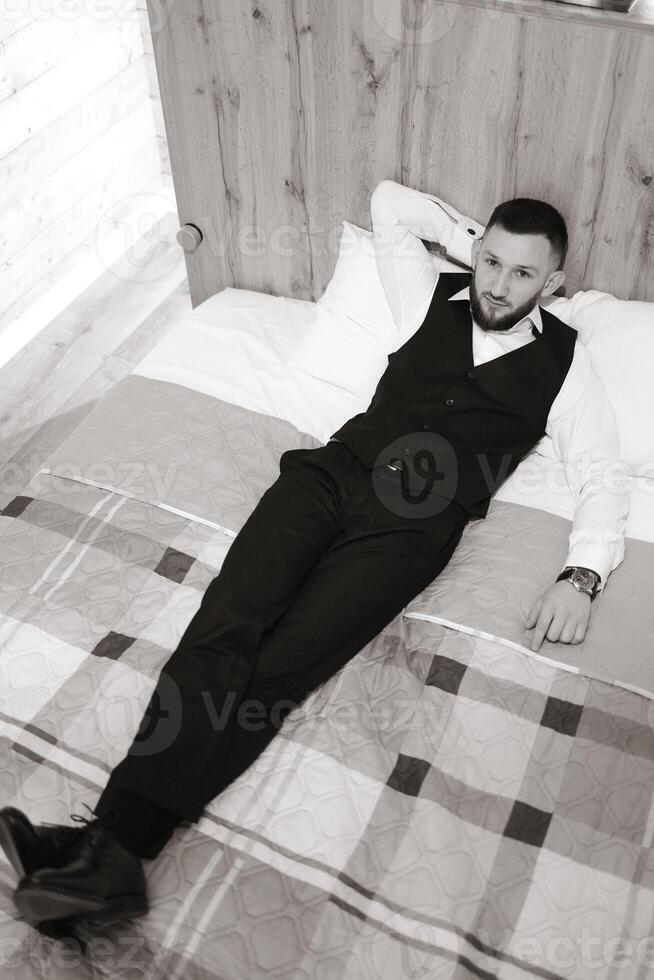 A man in a black vest and dress pants is laying on a bed. He is wearing a watch and has a beard photo