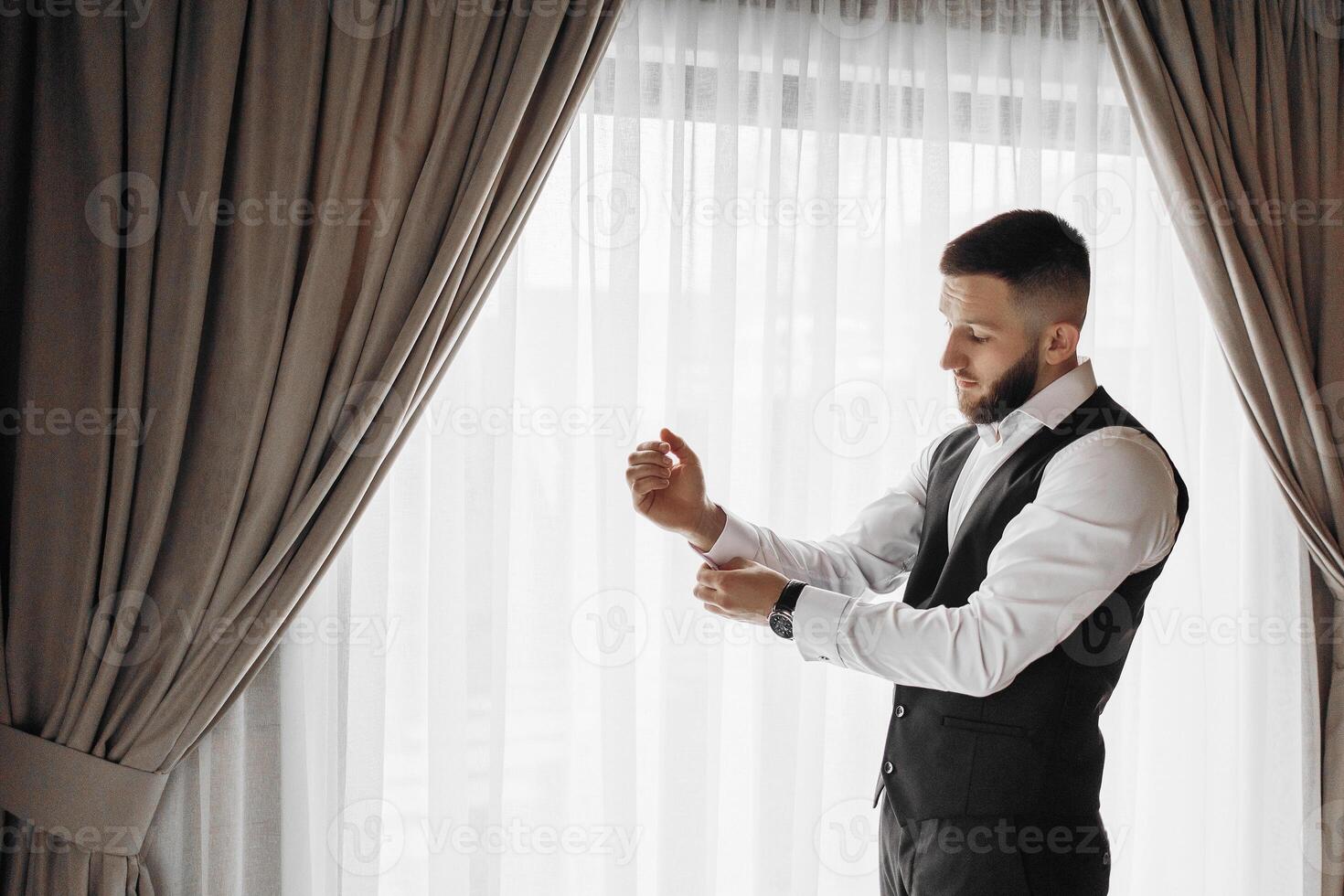 A man in a black vest and white shirt is getting dressed. He is wearing a watch and is adjusting his tie photo