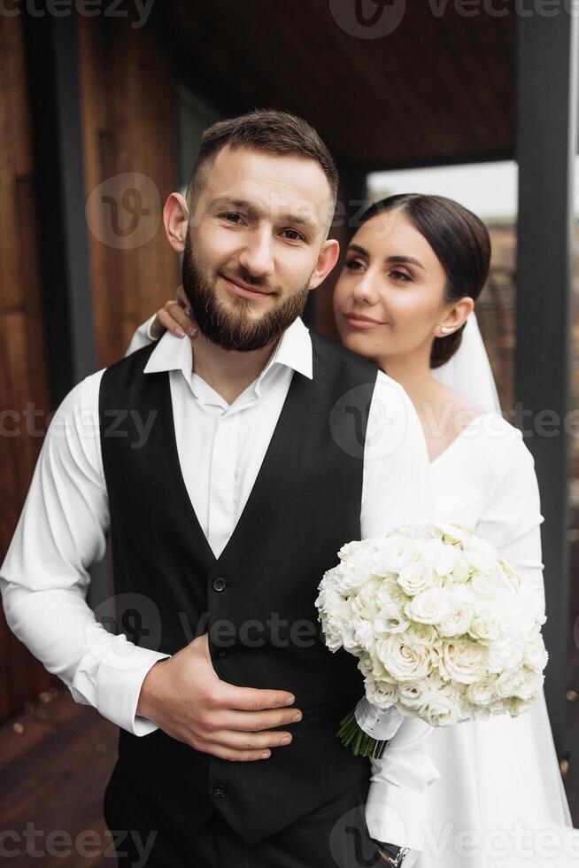 A man and woman are posing for a picture, with the man wearing a black vest and the woman holding a bouquet of white flowers. Scene is happy and celebratory, as the couple is likely getting married photo