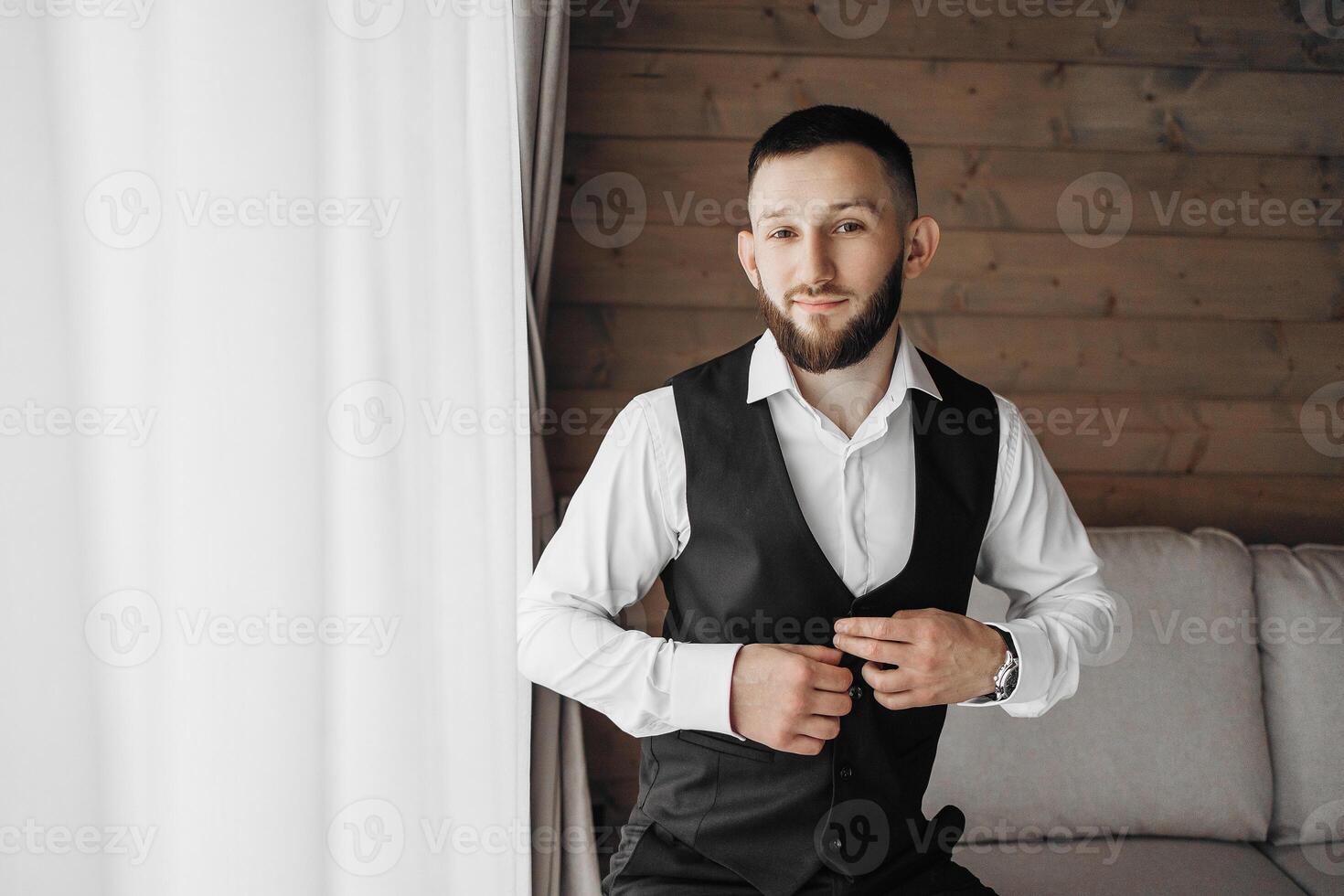 A man in a suit and vest is sitting on a couch. He is adjusting his vest and looking at the camera photo