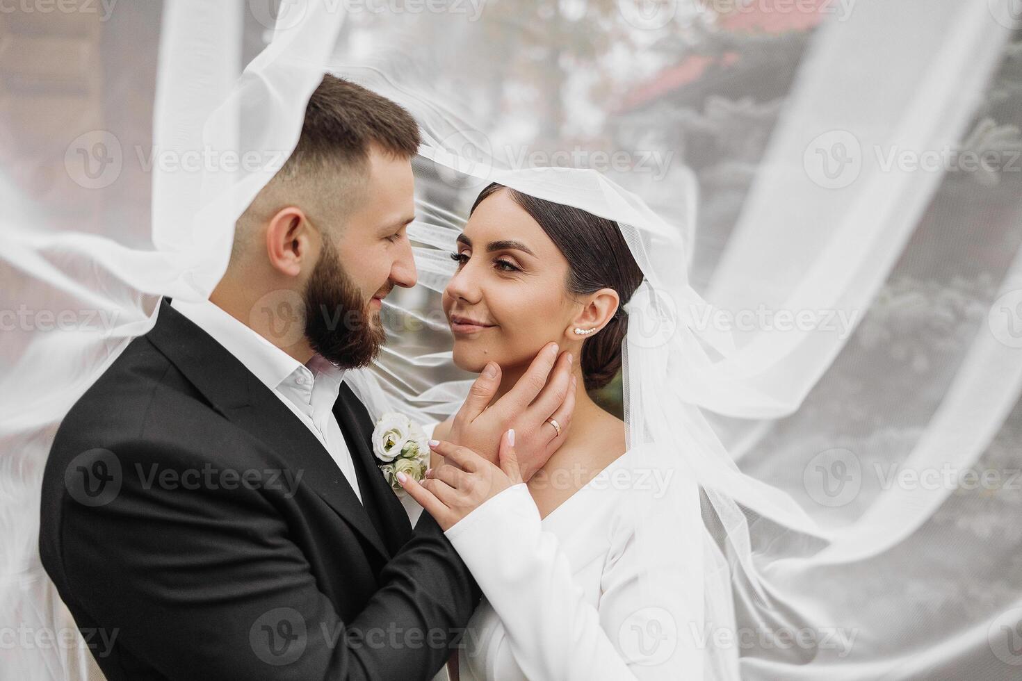 A bride and groom are standing under a white veil. The bride is wearing a white dress and the groom is wearing a black suit. They are both smiling and looking at each other photo