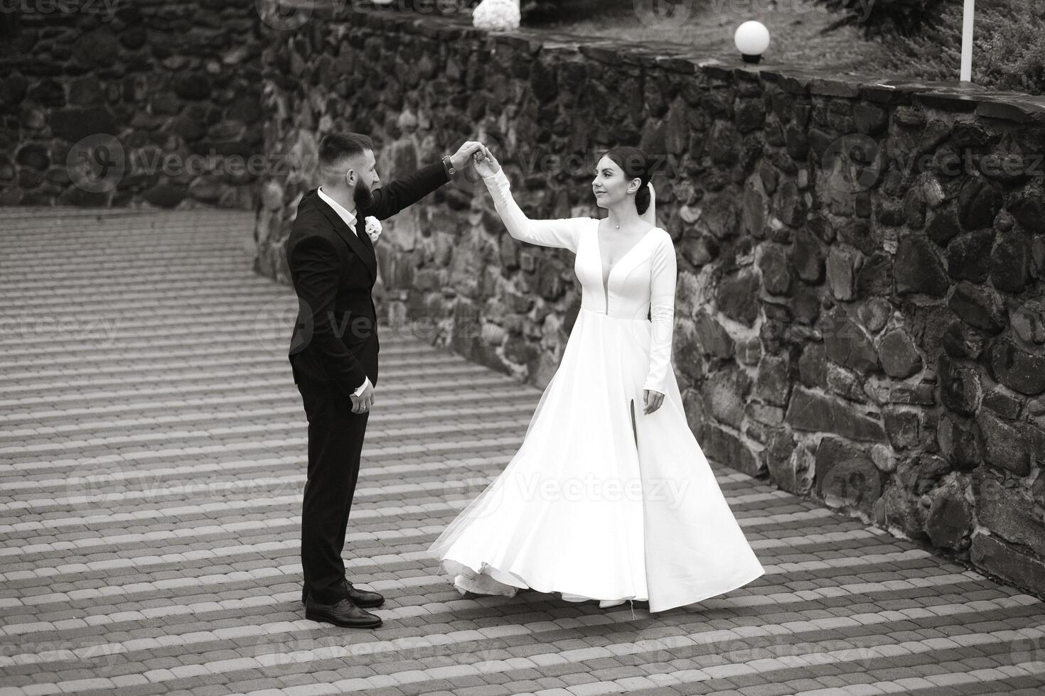 A bride and groom are dancing on a stone walkway. The bride is wearing a long white dress and the groom is wearing a suit. Scene is joyful and celebratory, as the couple is enjoying their wedding day photo