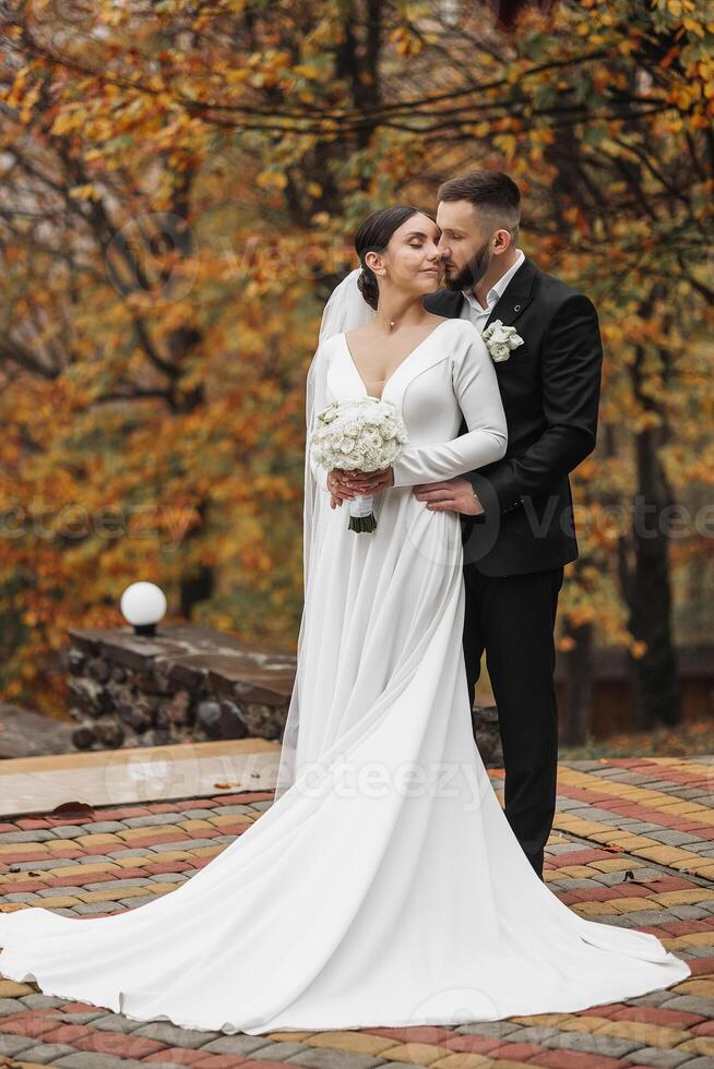 A bride and groom are posing for a picture in front of a tree. The bride is wearing a white dress and holding a bouquet, while the groom is wearing a suit. Concept of love and happiness photo
