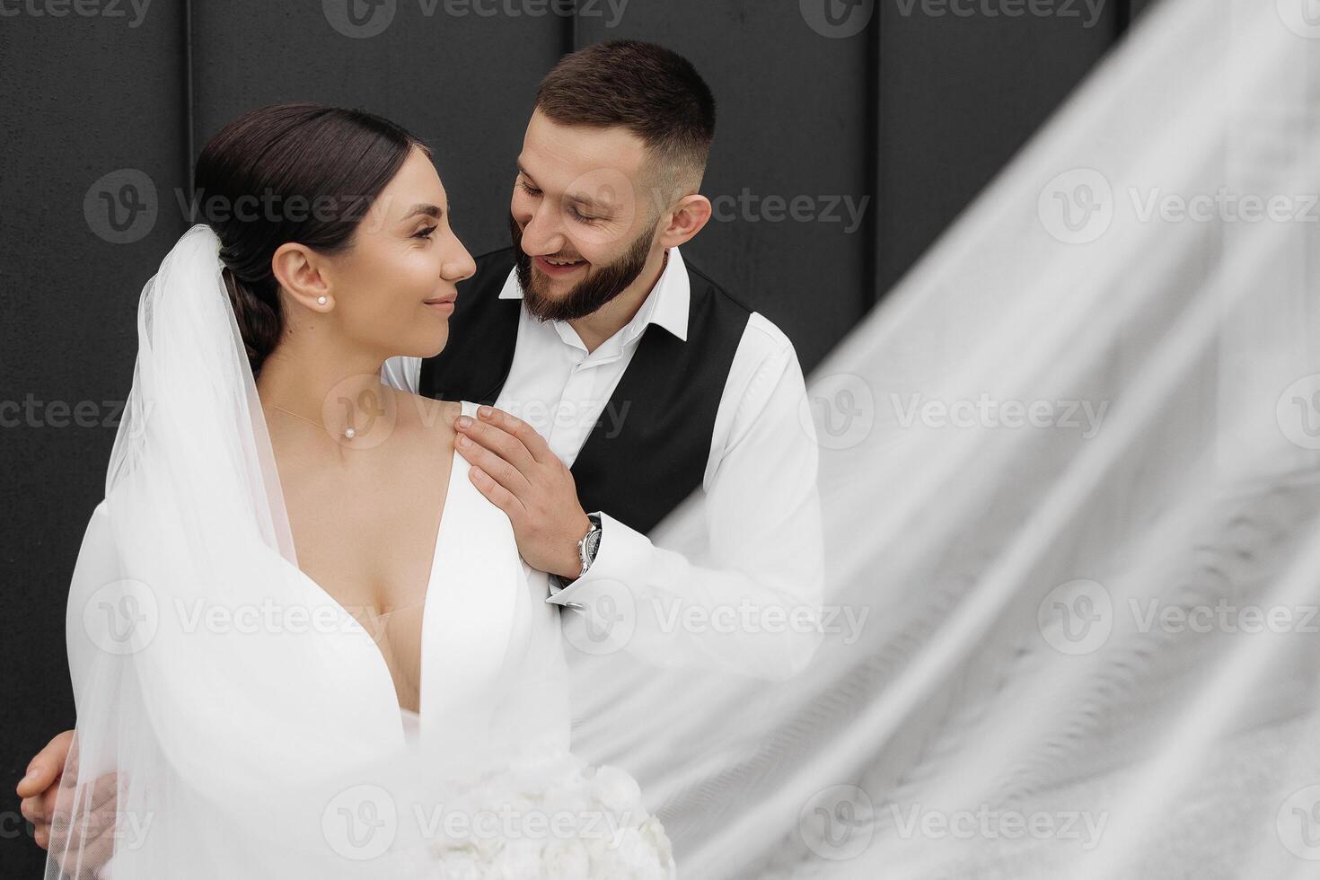 A bride and groom are posing for a picture in front of a white veil. The bride is wearing a white dress and the groom is wearing a black vest. Scene is happy and romantic photo