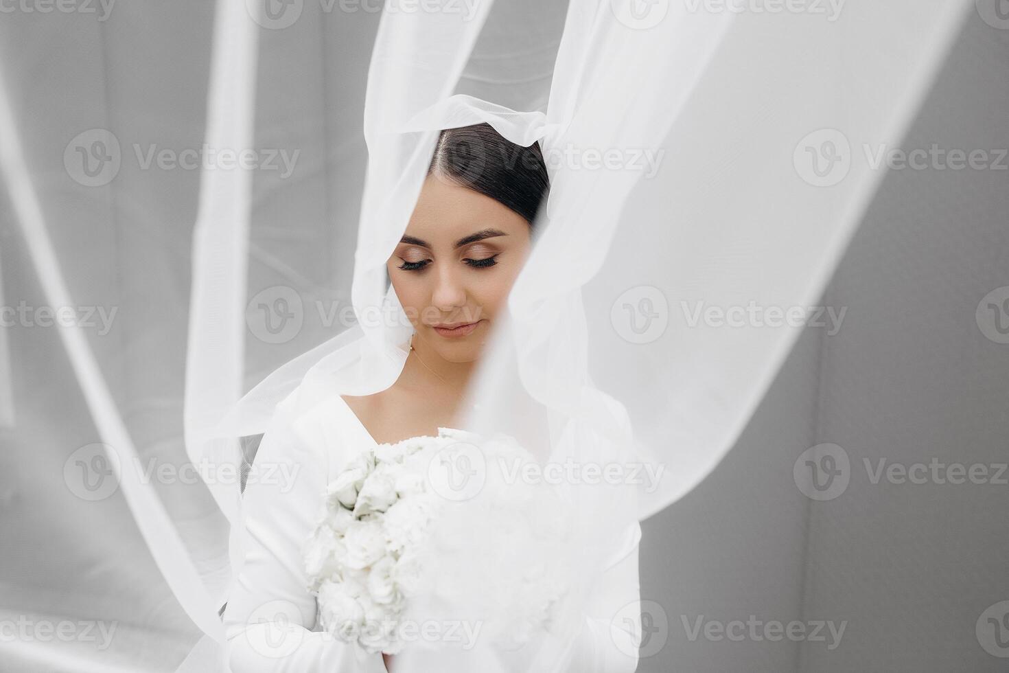 A woman is wearing a white dress and holding a bouquet of flowers. She is standing in front of a white curtain, which is partially covering her. Concept of intimacy and romance photo