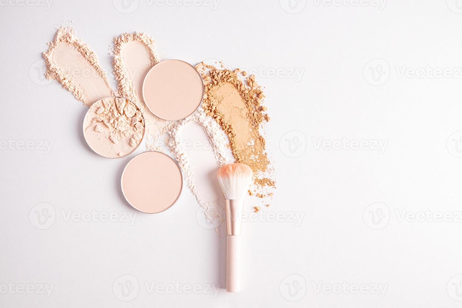 A few round blocks of compact powder in light tones and swatches of loose powder in different tones on a white background with a makeup brush photo