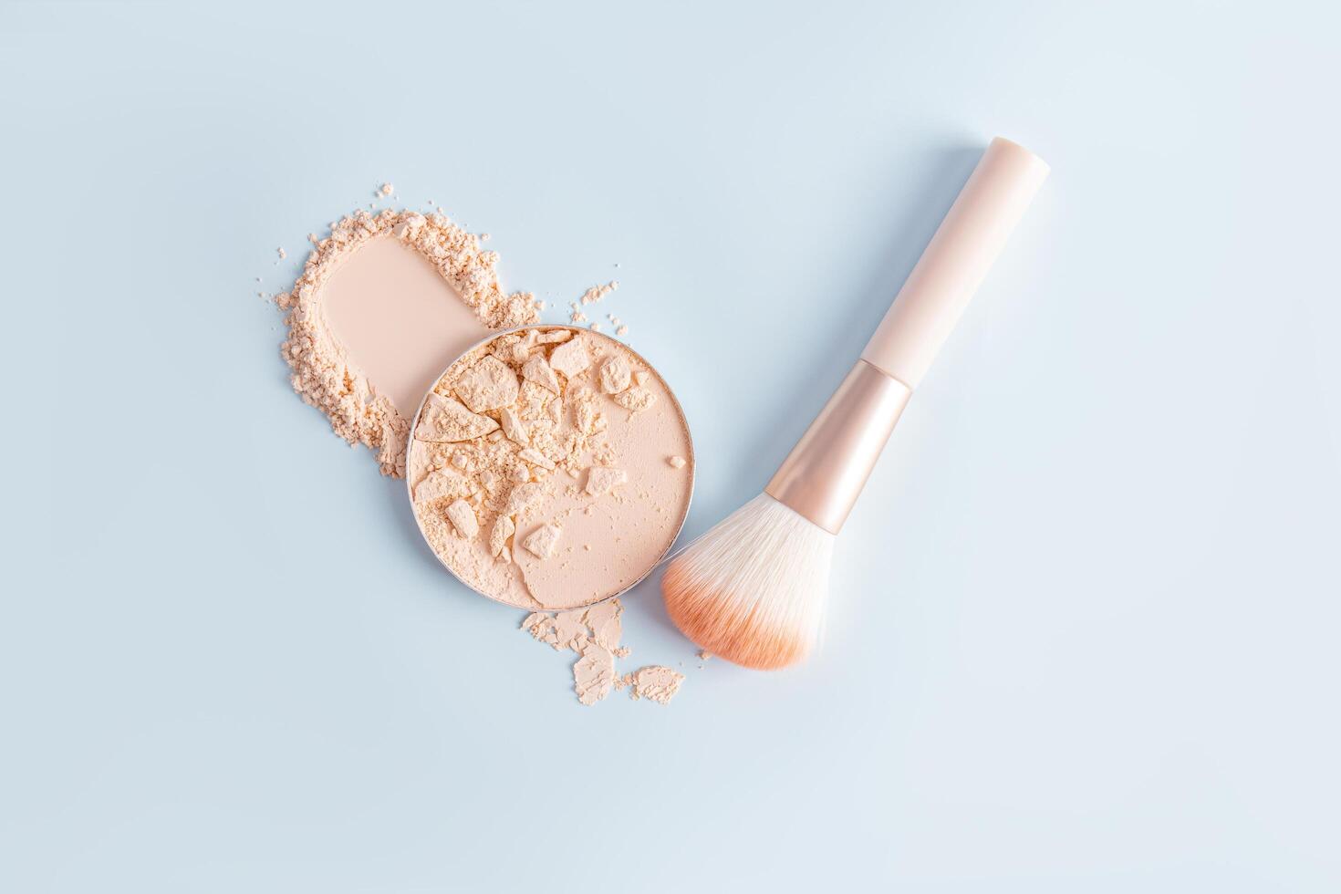 A round block of compact powder with broken pieces stands on a sample of loose powder of a natural tone with a makeup brush. Blue background. photo