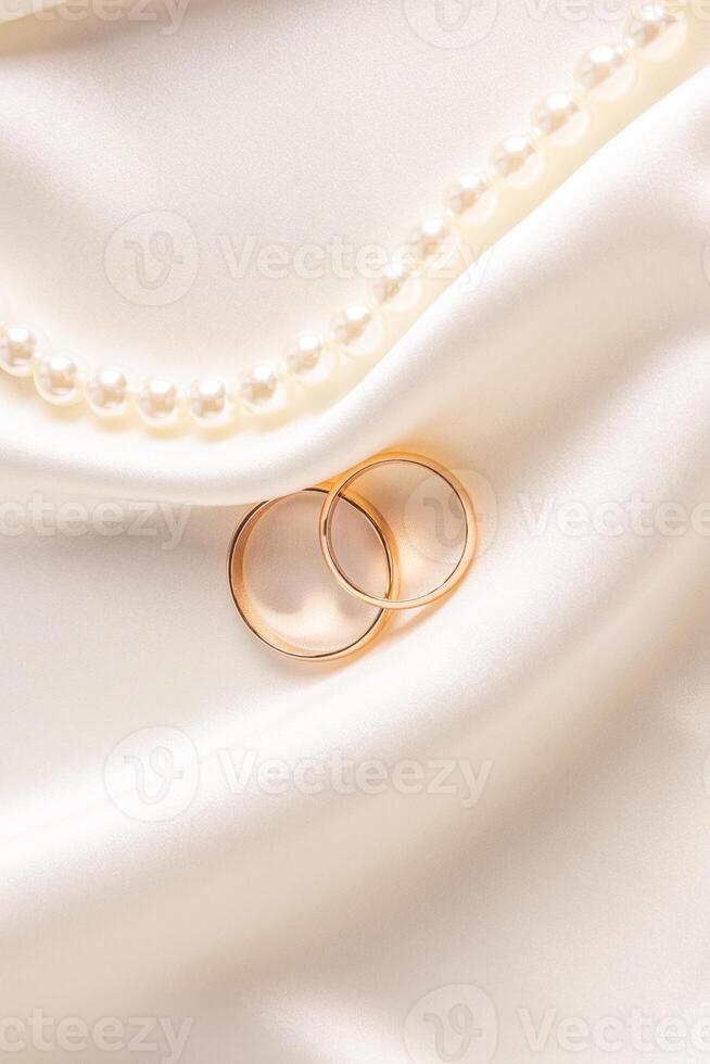 Two classic gold wedding rings lie among the delicate folds of satin fabric with a string of pearl beads. vertical front view. postcard, invitation. photo