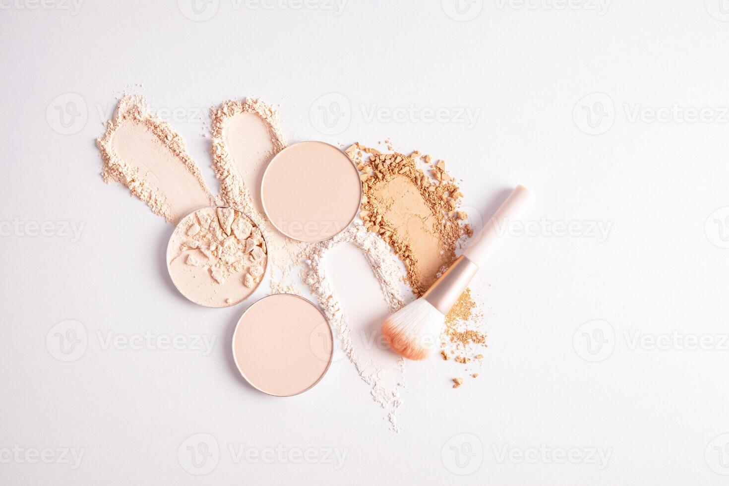 several round blocks of compact powder in light tones and swatches of loose powder in different tones on a white background with a makeup brush photo