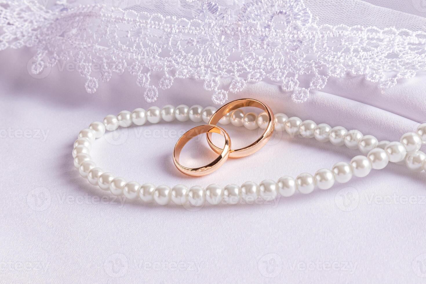 Two gold wedding rings lie on a string of pearl beads. on a white satin background with an embroidered veil. The concept of the wedding. Front view. photo