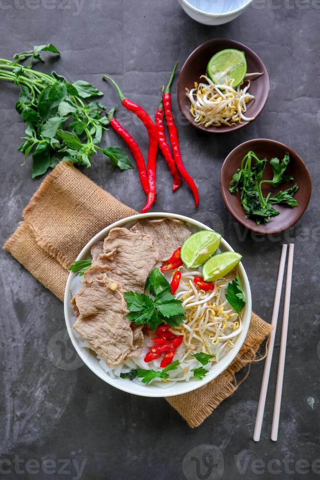 A Bowl Pho Bo traditional Soup with beef, rice noodles, ginger, lime, chili pepper in bowl. Close up. Vietnamese cuisine served on black background photo