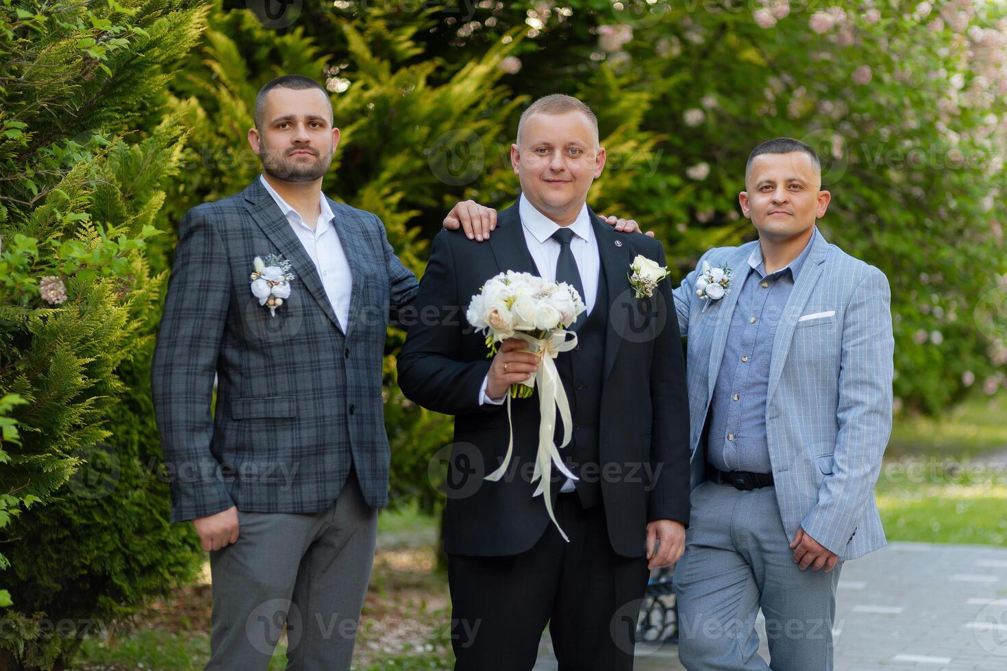 Three men are standing together, one of them holding a bouquet. They are dressed in suits and ties, and the man with the bouquet is wearing a tie as well photo