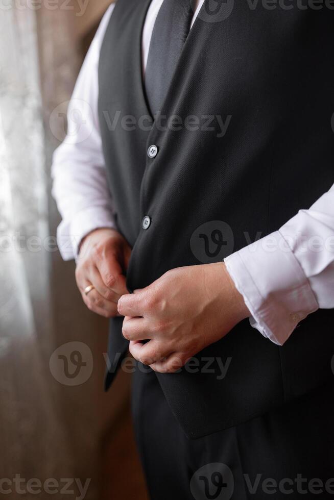 A man in a black vest and a white shirt is getting ready to dress up. He is adjusting his tie and putting on a ring. Concept of formality and preparation for a special occasion photo