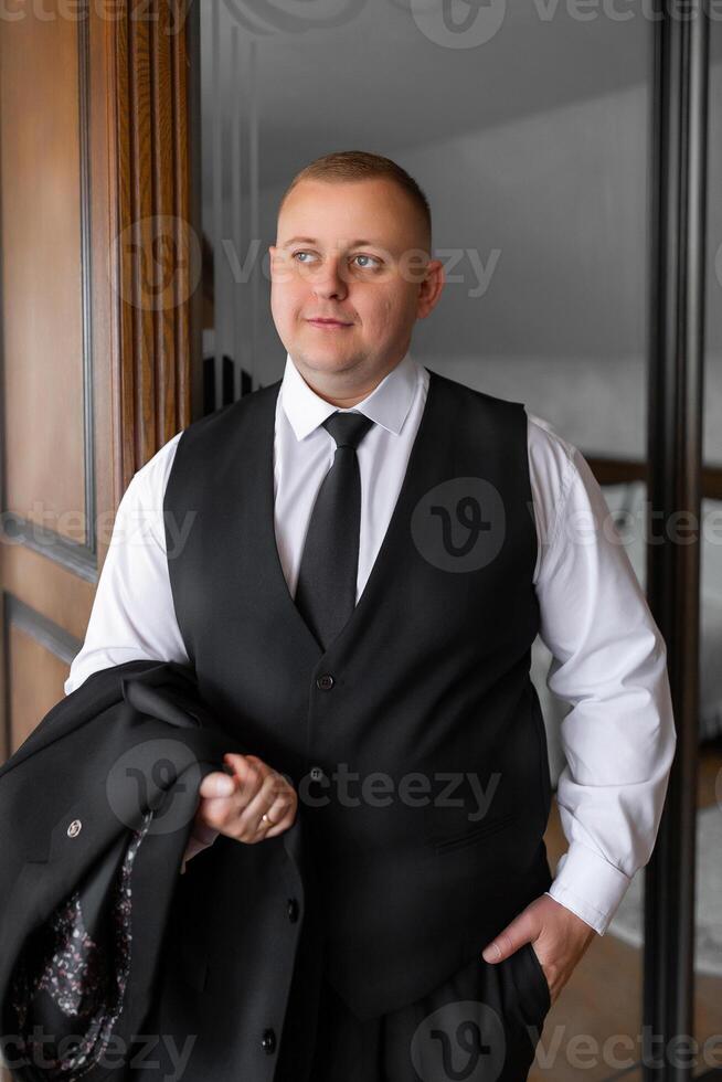 A man in a suit and vest stands in front of a mirror. He is wearing a black vest and a white shirt photo