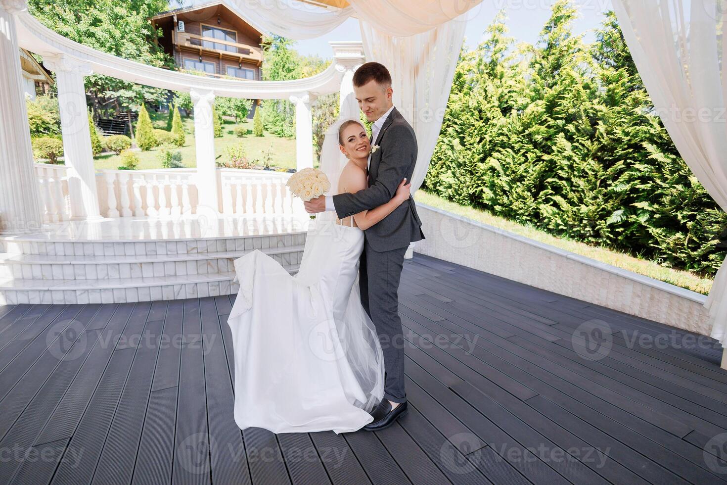 A bride and groom are standing on a wooden deck, with the bride holding a bouquet. Scene is happy and romantic, as the couple is celebrating their wedding photo