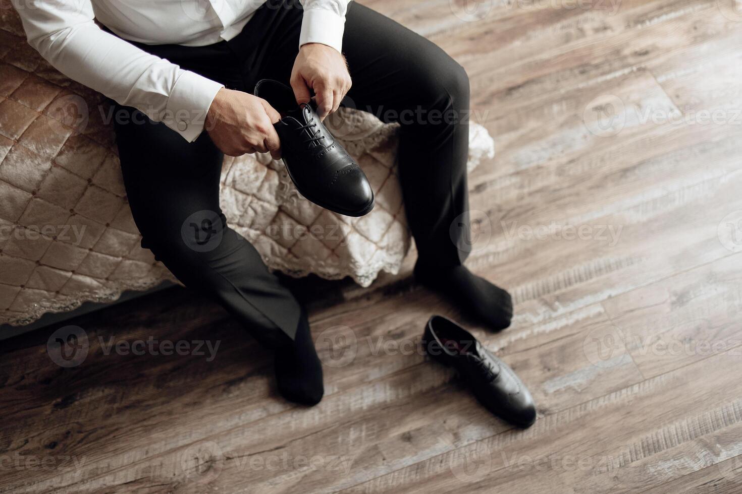 A man is sitting on a bed and putting on his shoes. The shoes are black and the man is wearing black pants photo