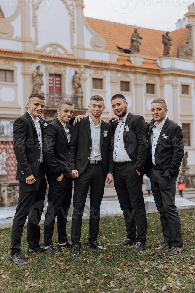 A group of five men in suits are posing for a photo in front of a building. Scene is formal and professional, as the men are dressed in suits and ties. The setting of the photo is a courtyard or park