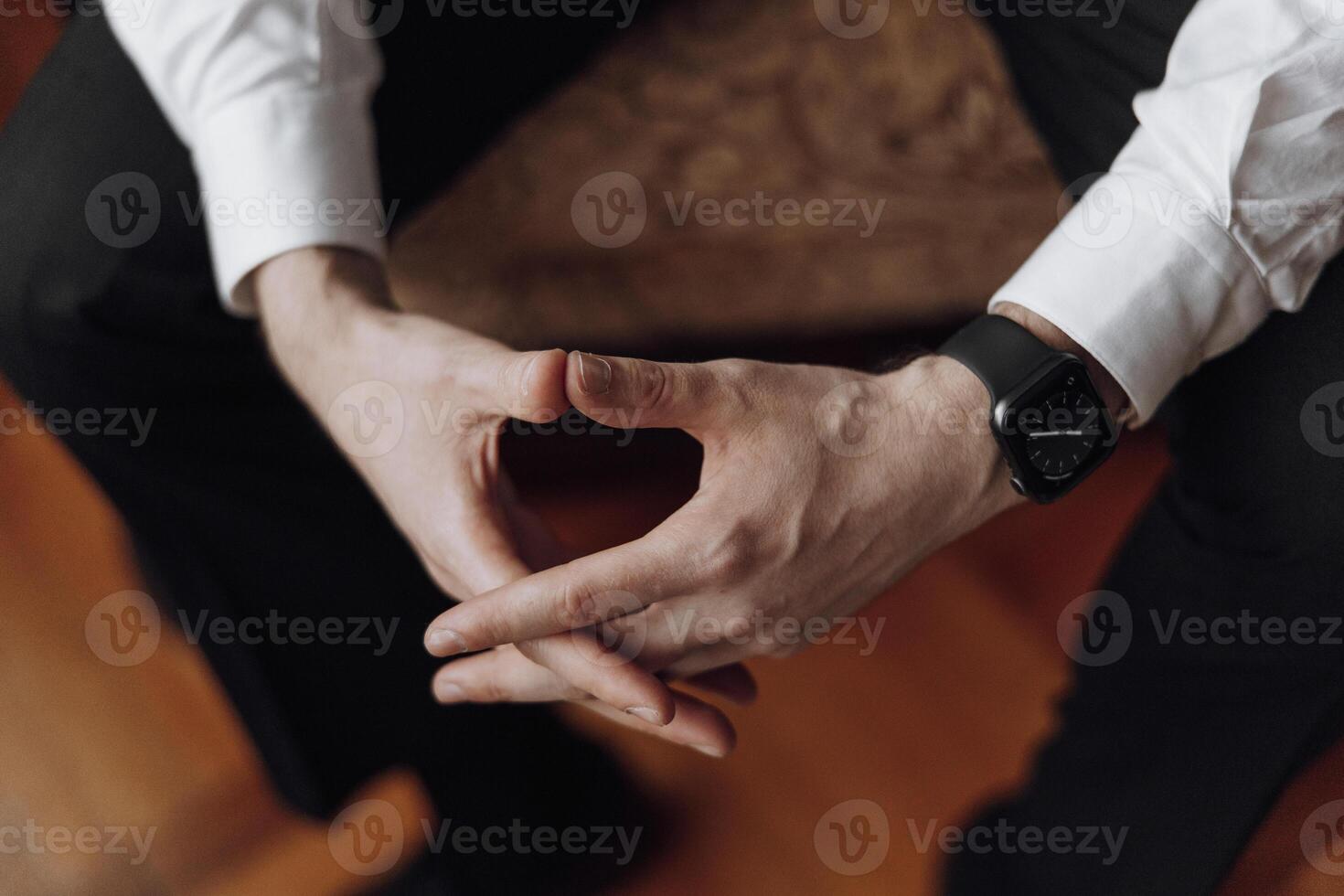 A man is sitting on a chair with his hands folded in his lap. He is wearing a watch and a white shirt. Concept of calmness and relaxation photo