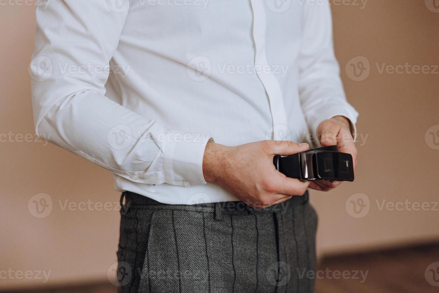 A man is wearing a white shirt and holding a belt. The man is wearing a grey plaid pants and a black belt photo
