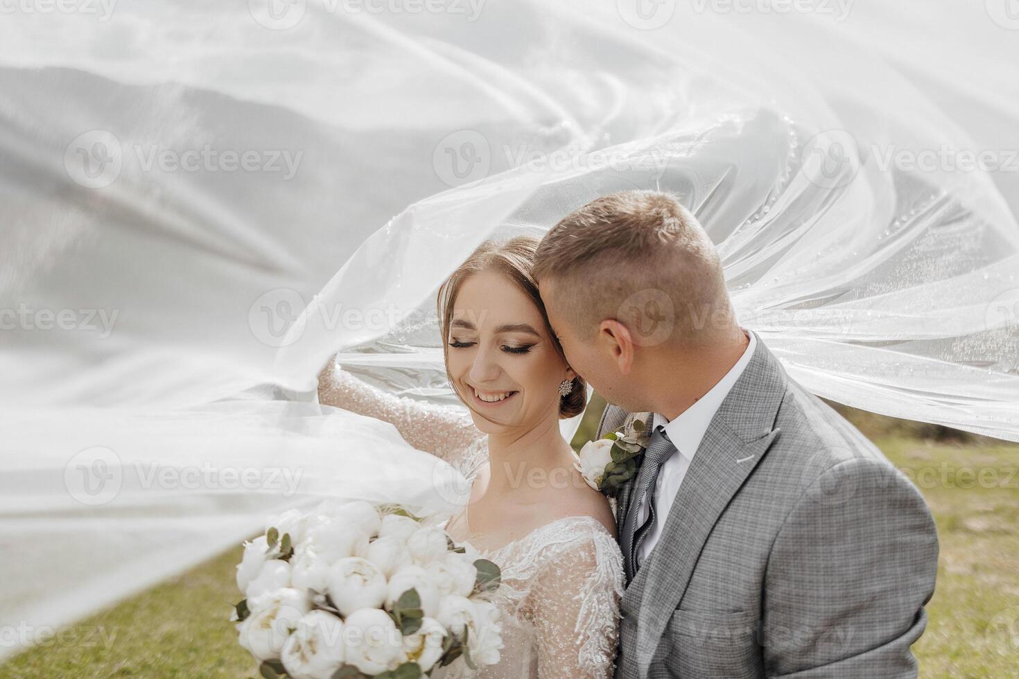 A bride and groom are standing in front of a white veil, with the bride holding a bouquet of flowers. Scene is romantic and happy, as the couple is embracing and enjoying their special day together photo