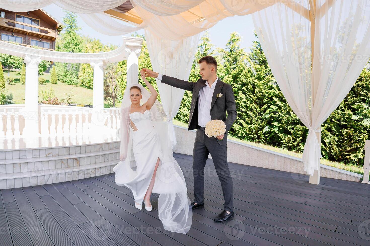 A bride and groom are dancing together in a garden. The bride is wearing a white dress and the groom is wearing a suit. They are both smiling and enjoying the moment photo