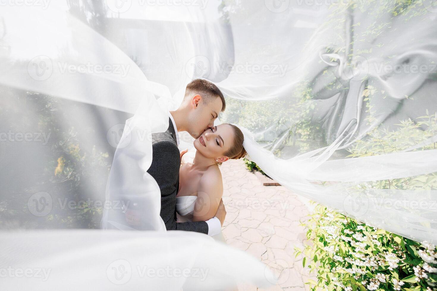 A bride and groom are kissing in front of a white veil. The bride is wearing a white dress and the groom is wearing a suit. The scene is romantic and intimate photo