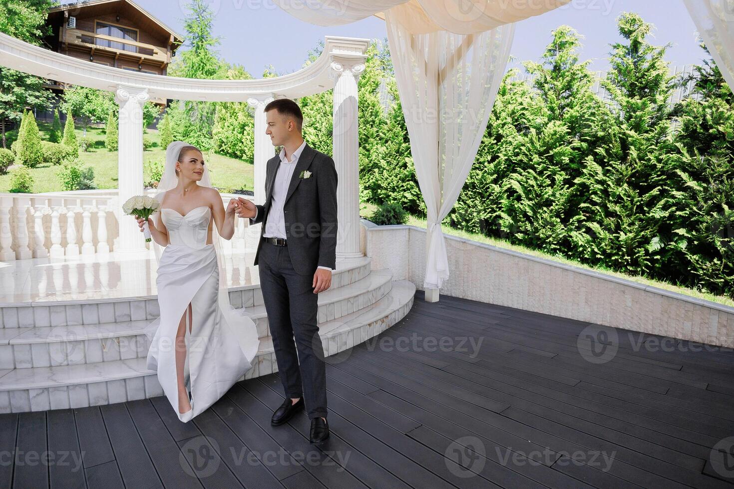 A bride and groom stand in front of a white archway. The bride is holding a bouquet and the groom is holding her hand photo