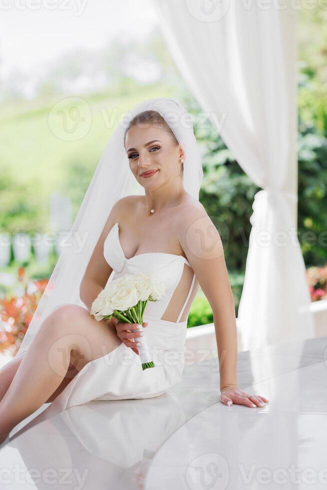 A bride is sitting on a table with a bouquet of flowers in her lap. She is wearing a white dress and a veil. The scene is set in a garden or a courtyard, with a white curtain in the background photo