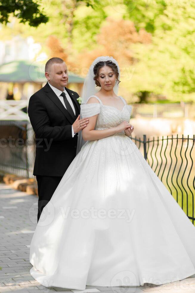 A bride and groom are posing for a picture in front of a fence. The bride is wearing a white dress and the groom is wearing a black suit. Scene is happy and celebratory photo